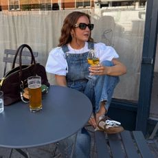 Influencer @rebeccaferrazwyatt sits at a table outfit wearing Miu Miu X New Blance backless trainers with dungarees and a puff-sleeve blouse.