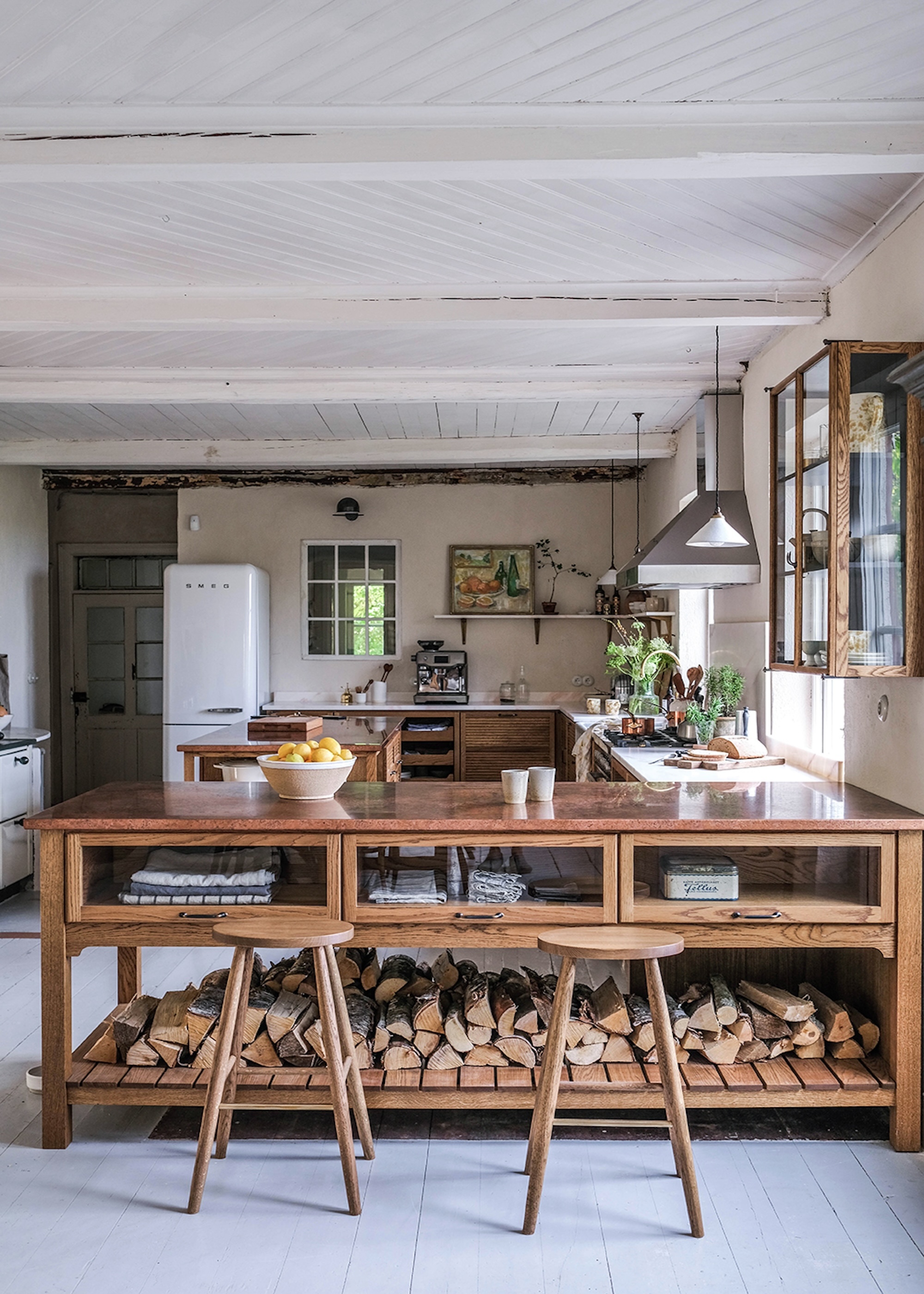 A view of the kitchen with a wooden and glass island with wooden stools in front. There is a barn-style exposed beam ceiling and cream colored walls.