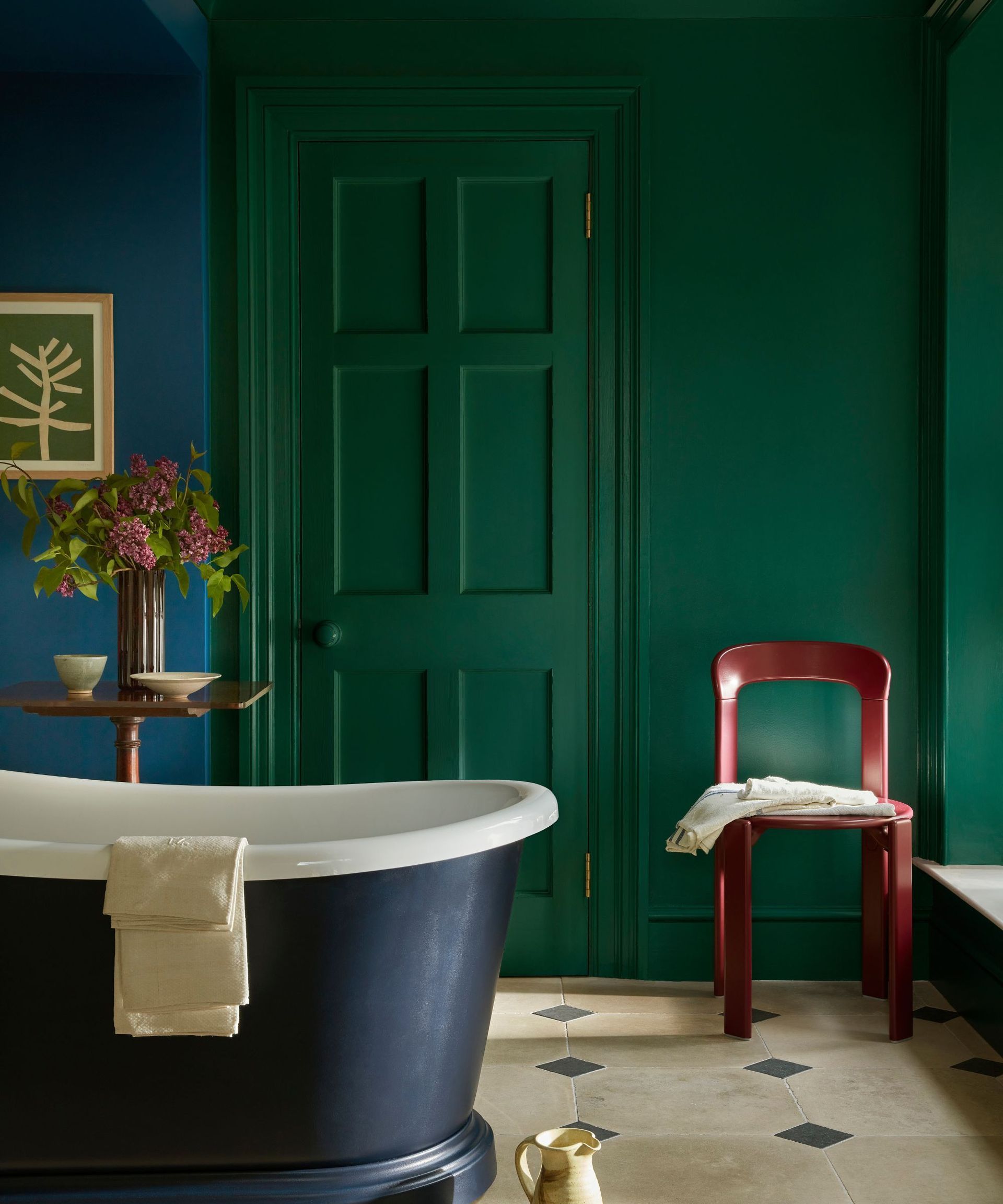 A bathroom with a freestanding roll top bath, a small brown wooden table beside it with decorative items on top. There is a red chair in the corner and classic cream and black tiles on the floor. The wall and the interior door has been painted using the color drenching method, in a vivid and highly pigmented dark green.