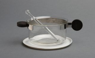 A glass stirrer in a glass sauce pan featuring black round handles which is place on a white round ceramic plate photographed against a grey background