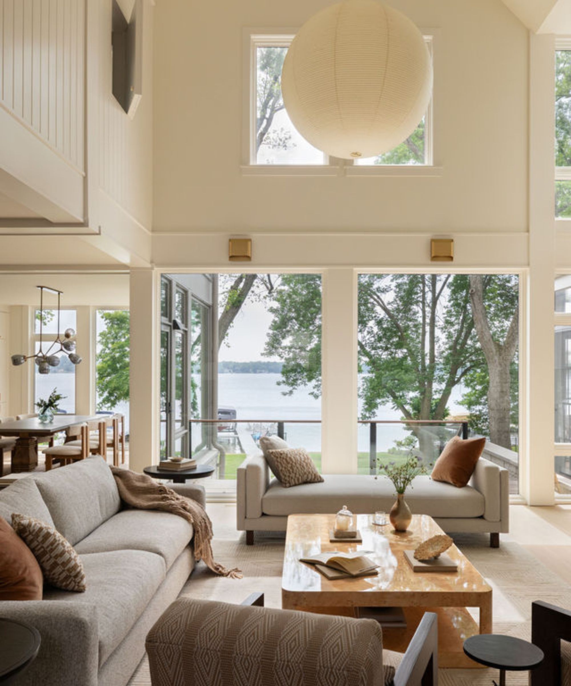 Large sunroom overlooking a lake with large windows, high ceilings, and a balcony. A grey sofa, a daybed and a burl wood coffee table at the centre, with a dining room in the background.