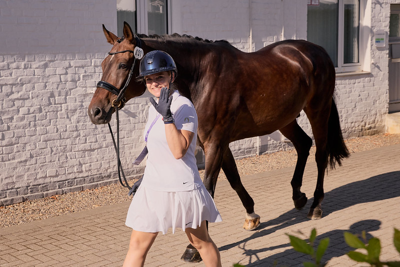 A lady wearing a white top, skirt and riding hat leads a dressage horse in a bridle on their way to the horse inspection in Aachen.