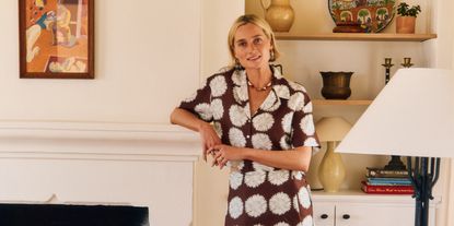 A young woman dressed in a patchworky, brown and white matching set with short sleeves stands against a fireplace in a layeredly decorated, sun-filled room.