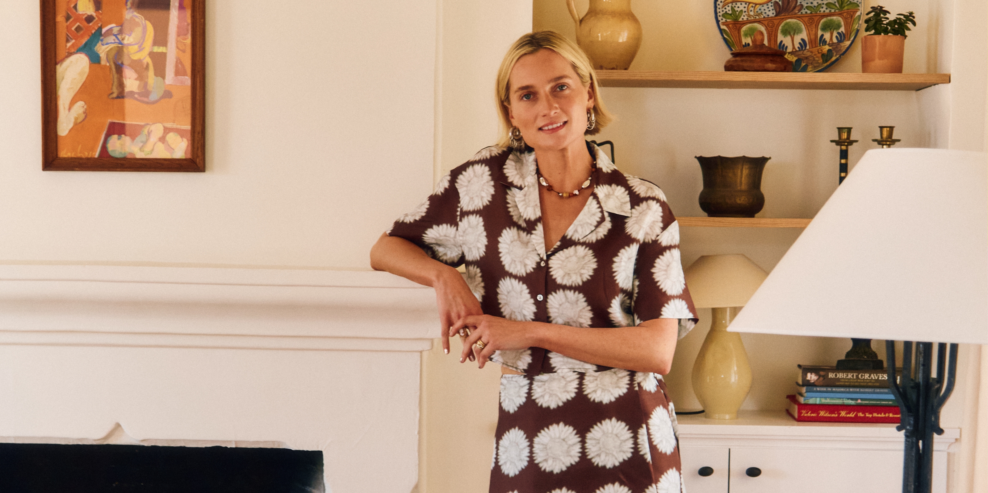 A young woman dressed in a patchworky, brown and white matching set with short sleeves stands against a fireplace in a layeredly decorated, sun-filled room.