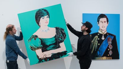 A man wearing black and one in blue hangs a turquoise pop art Andy Warhol painting of Princess Diana next to one of Prince Charles