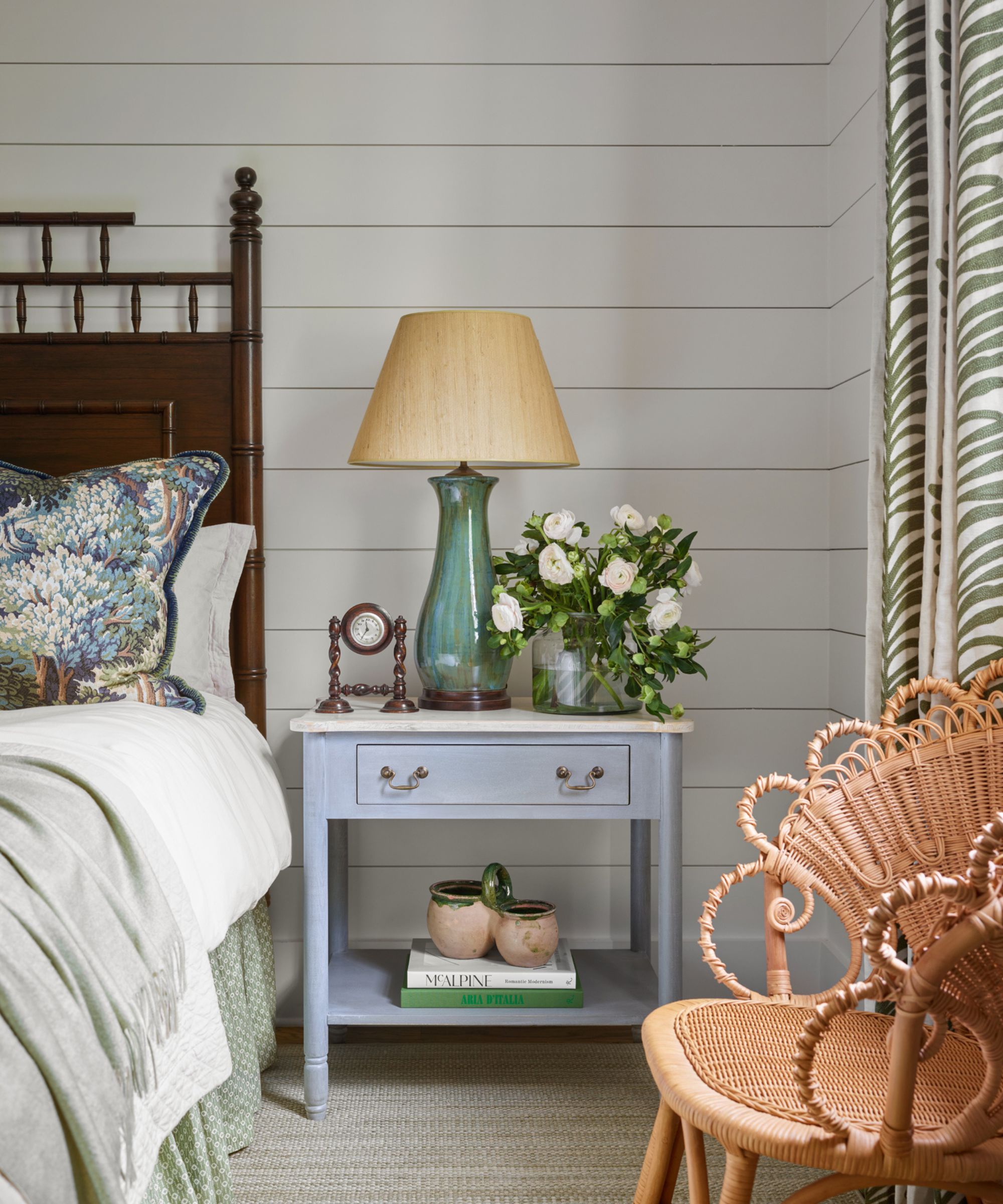 Bedroom with shiplap walls, a blue nightstand with a glass lamp and a vase of roses, a rattan armchair and a mahogany antique bedframe