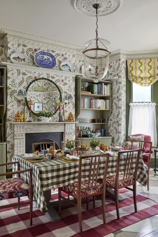 colorful dining room with stylised leaf wallpaper, green painted woodwork, check tablecloth and rug, vintage chairs with upholstered seats, painted shelving units, mirror, plates on wall, looks, mirror, blinds