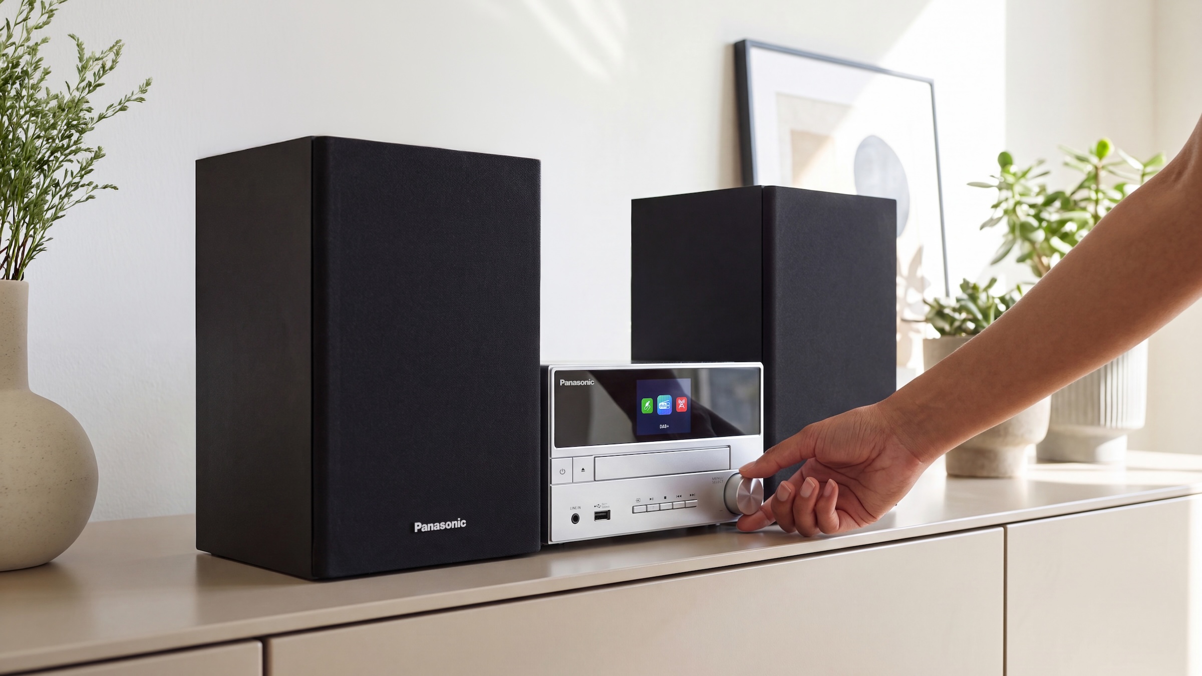A Panasonic SC-PM302 hi-fi system on a cabinet in a modern lounge with a hand adjusting the front dial.