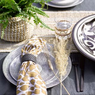 Place setting with rattan runner, pot plant in a woven pot, and monochrome plates topped with a animal print napkin