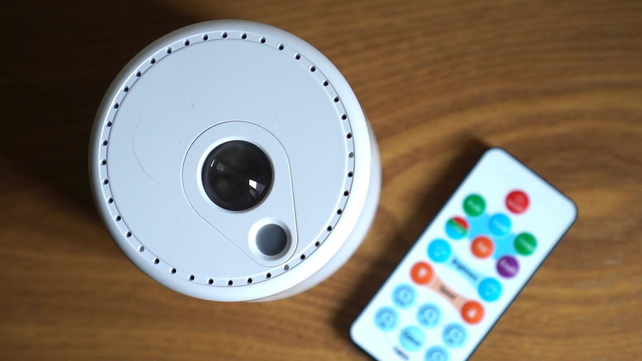 A white spherical projector is next to a multicolor remote control, both on a brown wooden surface.
