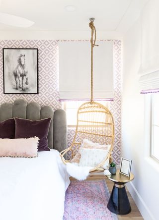 A bedroom with pink wallpapered walls, a bed with a gray headboard, and a wicker swing chair suspended in the window