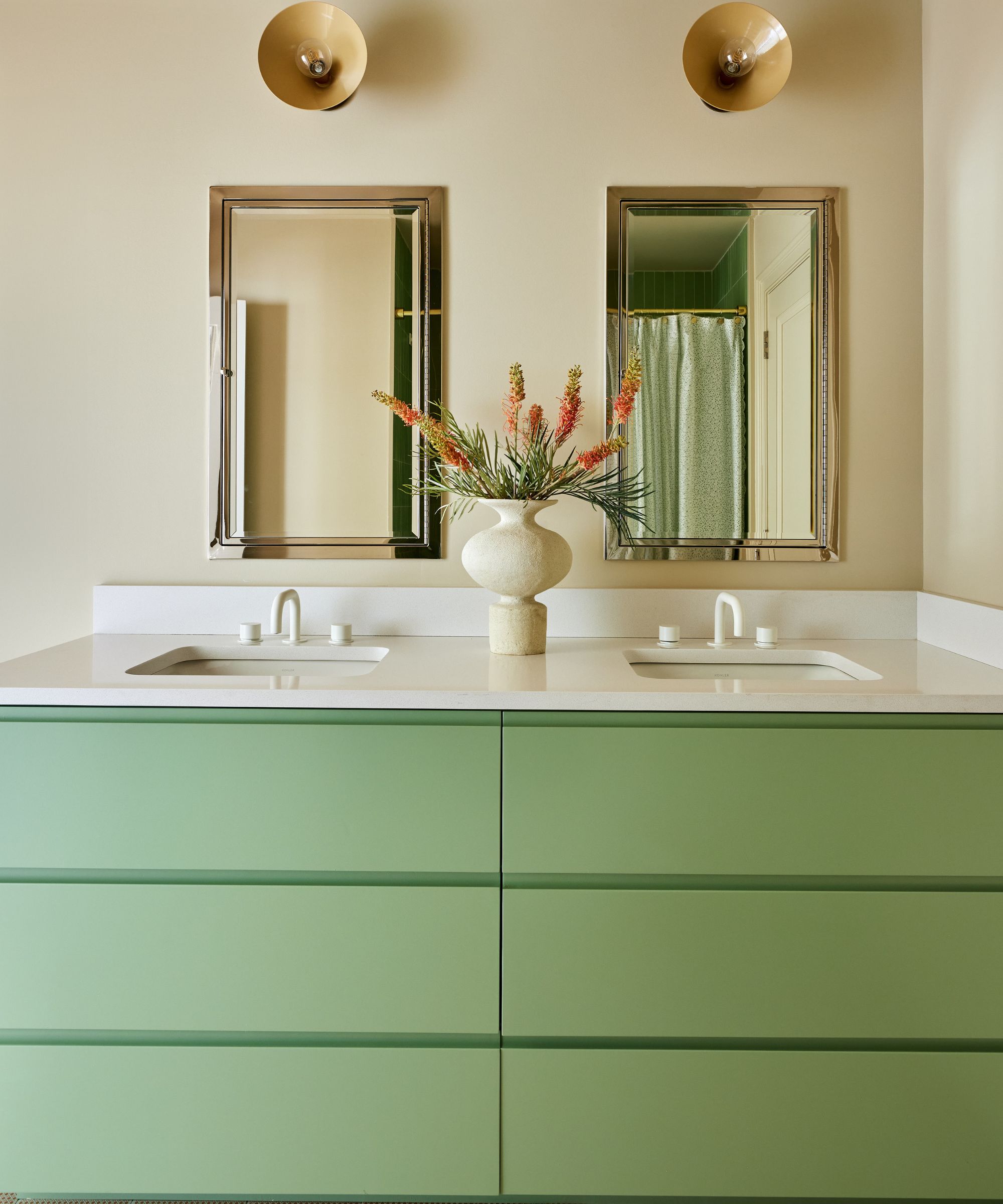 Double vanity unit with two sinks, two mirrors, round wall scocnes and green drawers and vase of flowers
