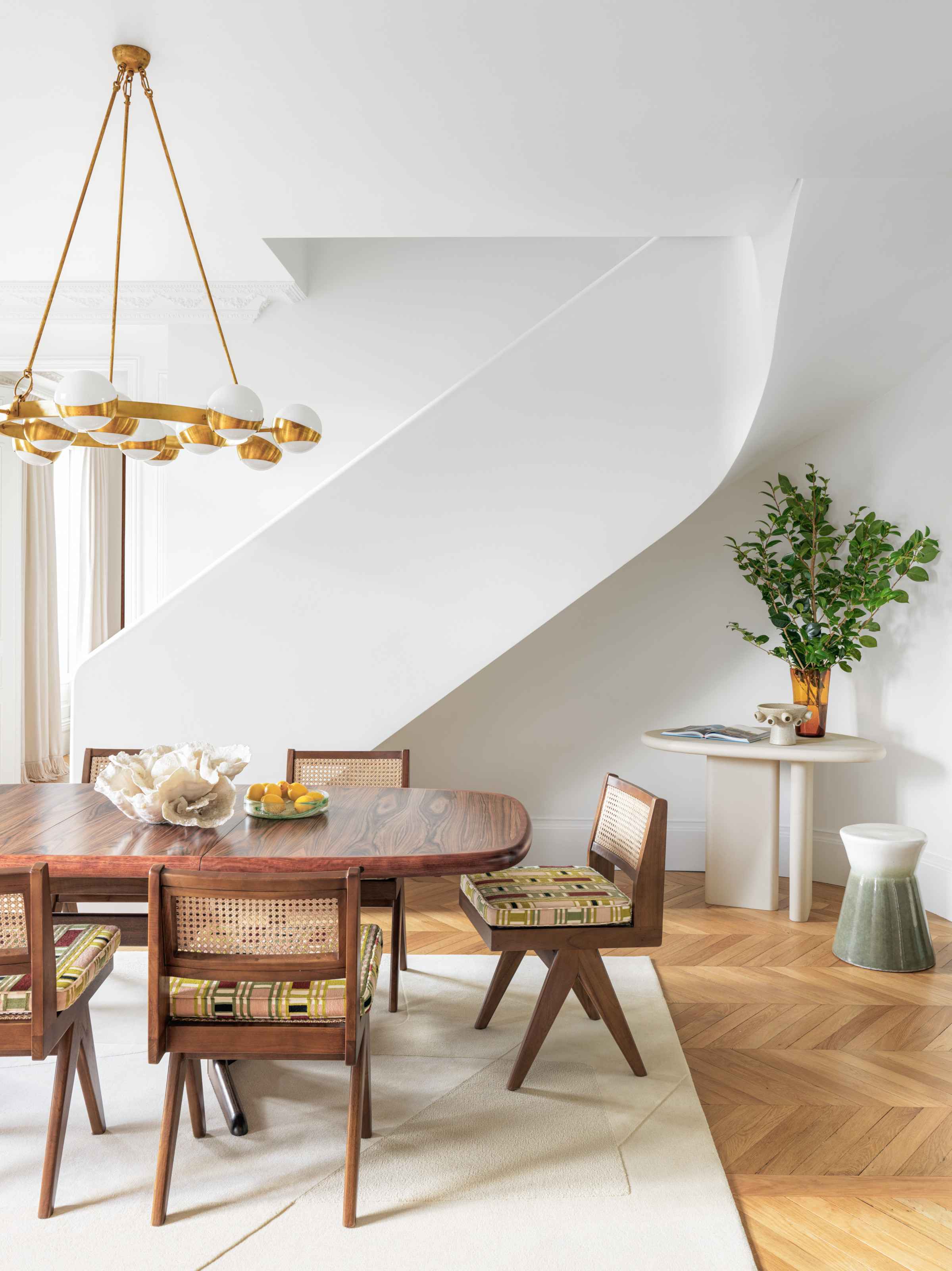 White dining room with curved staircase, wooden oval table and angular chairs, gold and white globe chandelier and white accent table