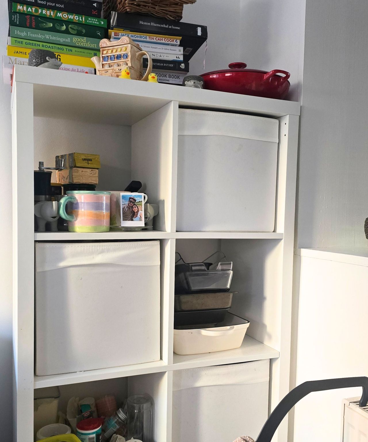 A white kitchen storage unit with books, a teapot, and a red kitchen pan on top of it, and mugs, trays, baking ingredients, and white storage cubes inside it
