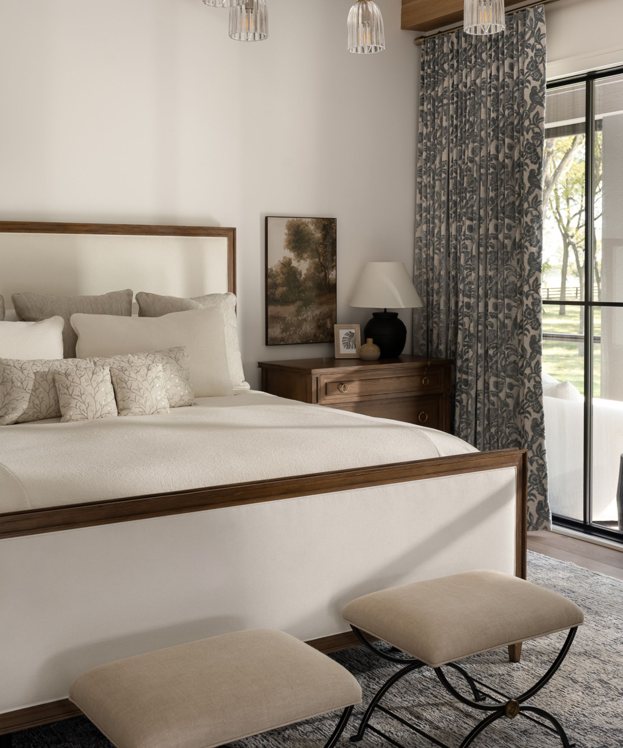 A serene bedroom featuring a wood-trimmed upholstered bed with neutral linens, a wooden nightstand with a black lamp, and blue patterned floor-to-ceiling curtains.