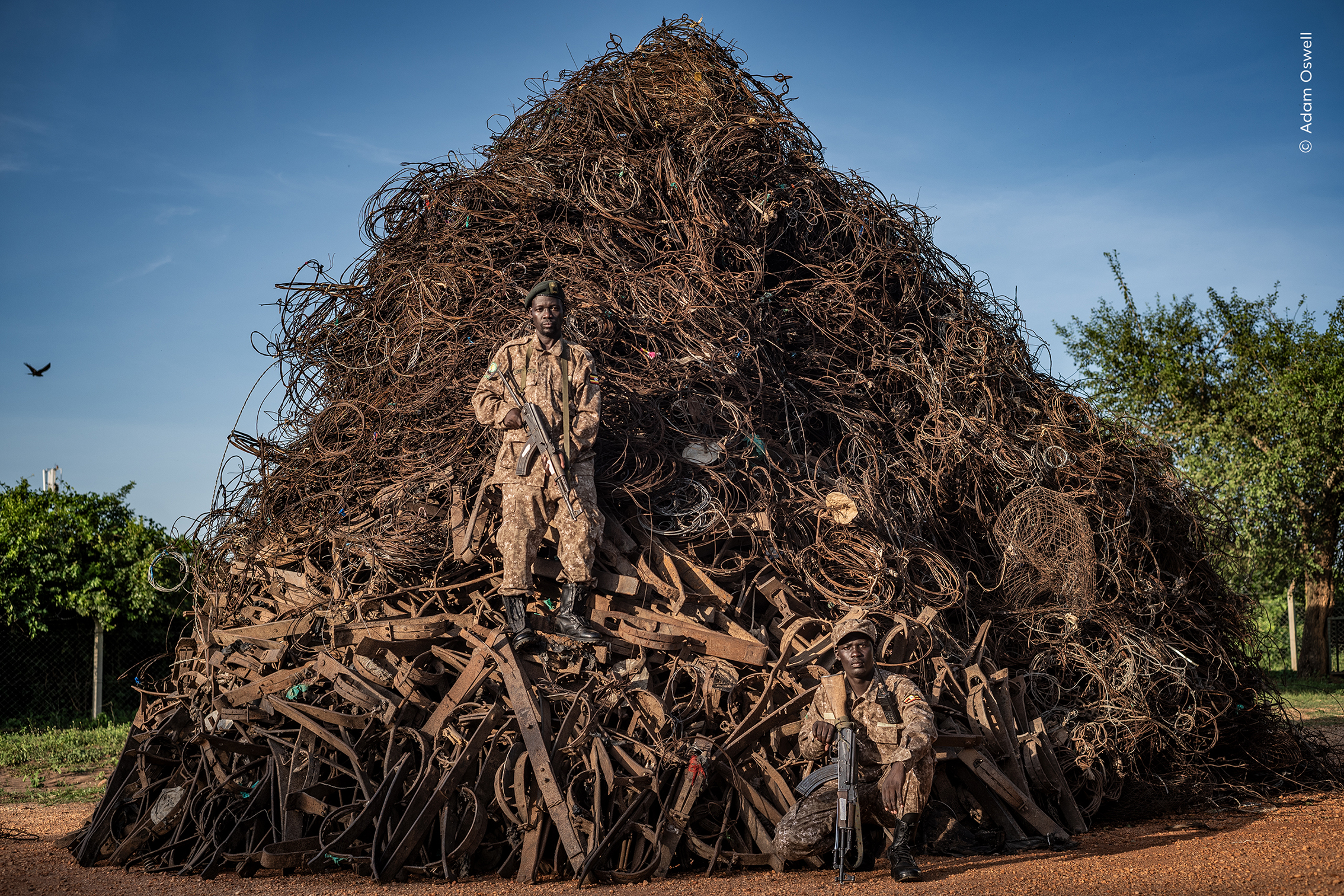 Two armed soldiers pose in front of a large, chaotic pile of twisted metal and scrap, set against a clear blue sky