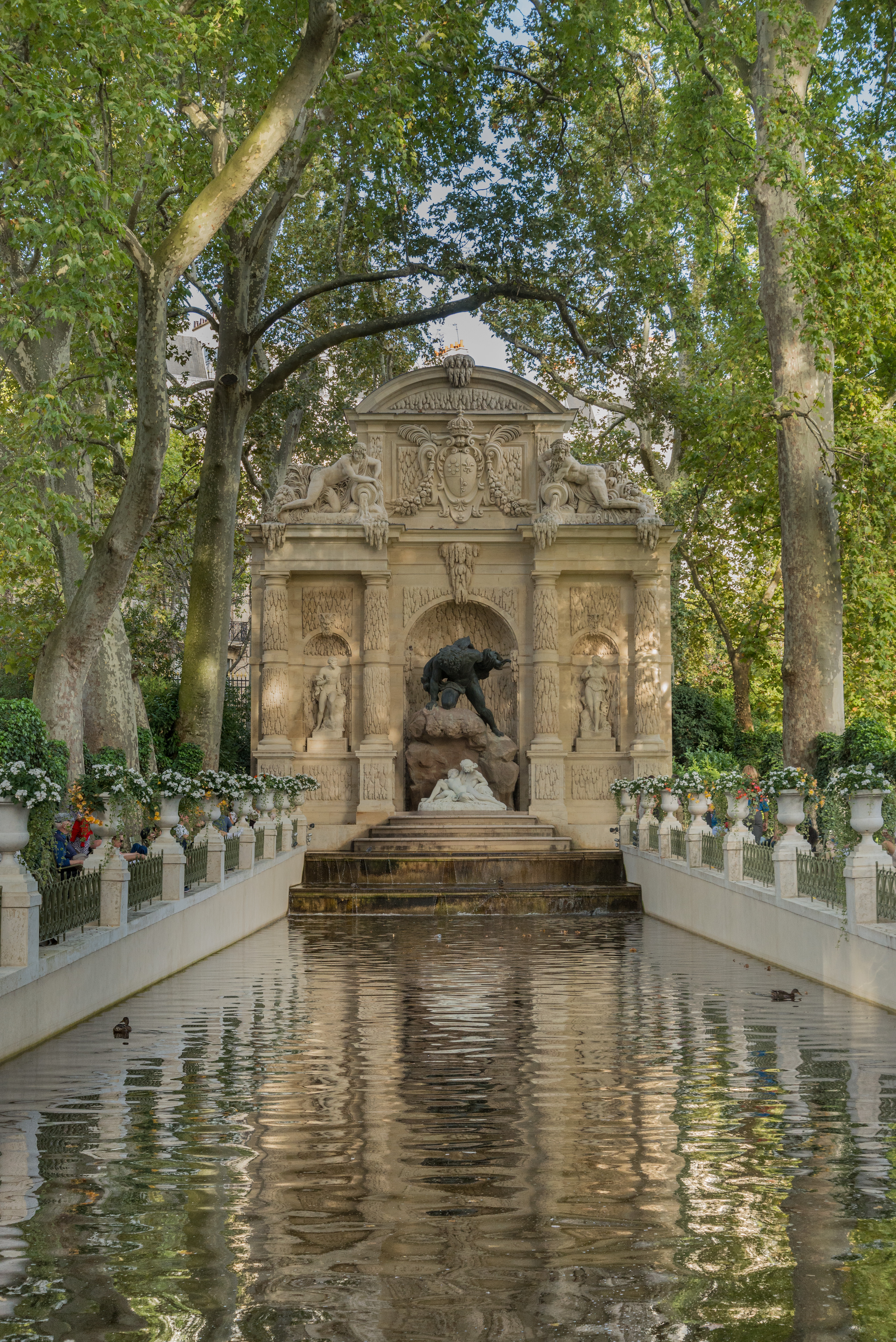 A picturesque garden view with a central fountain decorated with an ornate marble wall, potted plants, and thriving, sun-lit trees.