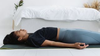 A woman is seen in a white bedroom lying on her back on the floor on a yoga mat, next to her bed