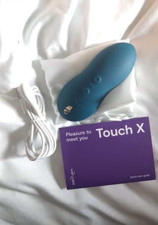 We-Vibe Touch X in dark blue with charging cable and instruction booklet on a white bed