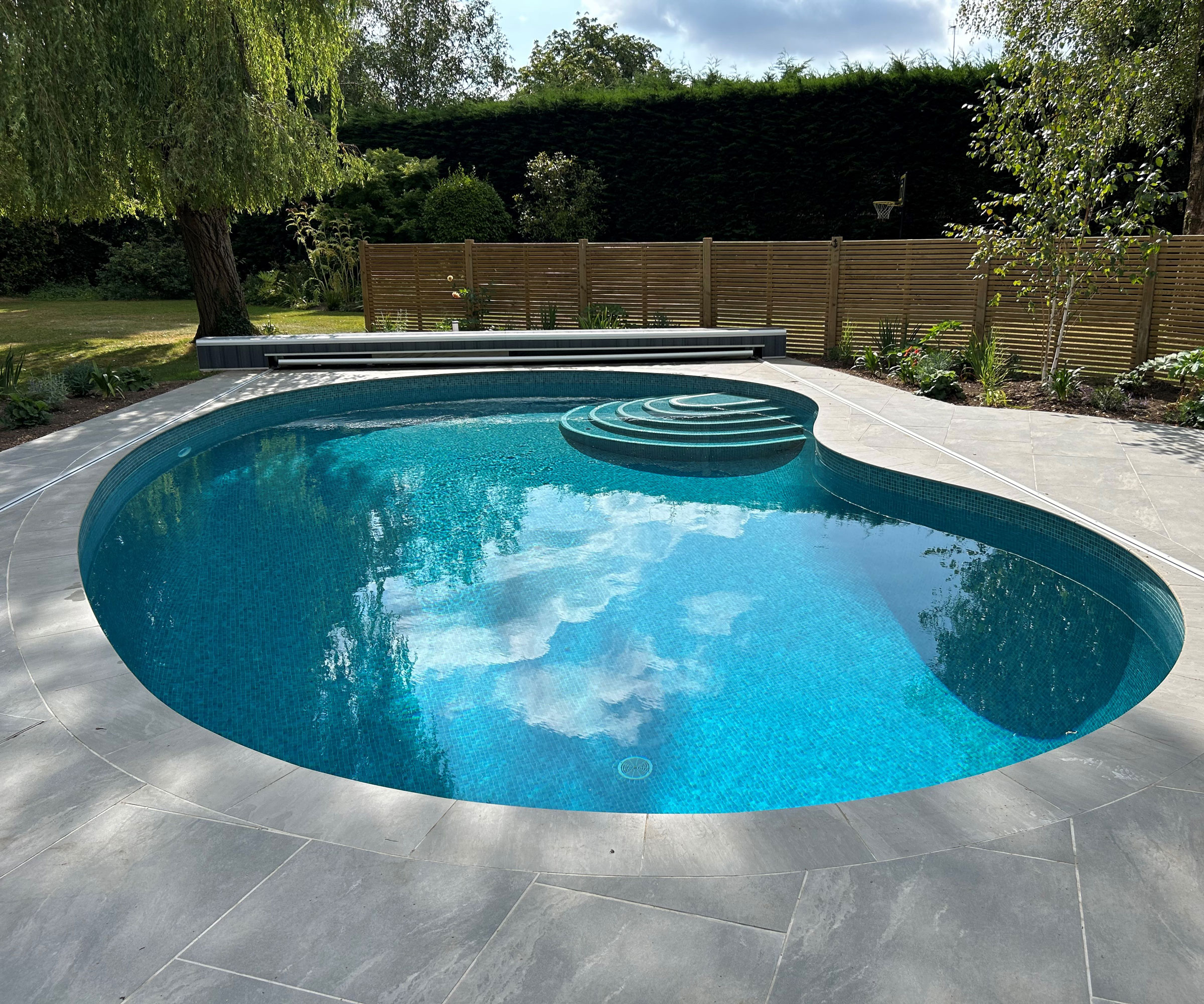 kidney shaped swimming pool with reflections of the sky in the water and trees offering areas of shade in the pool, surrounded by paving, a fence and planting