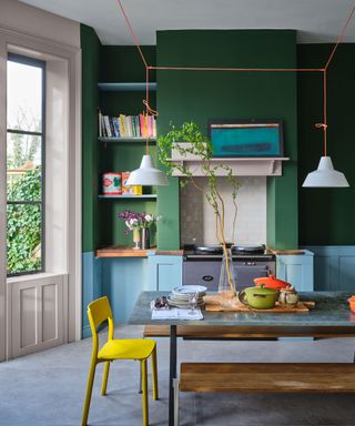 Green and blue kitchen with wooden dining table, gray flooring, low hanging pendant lights