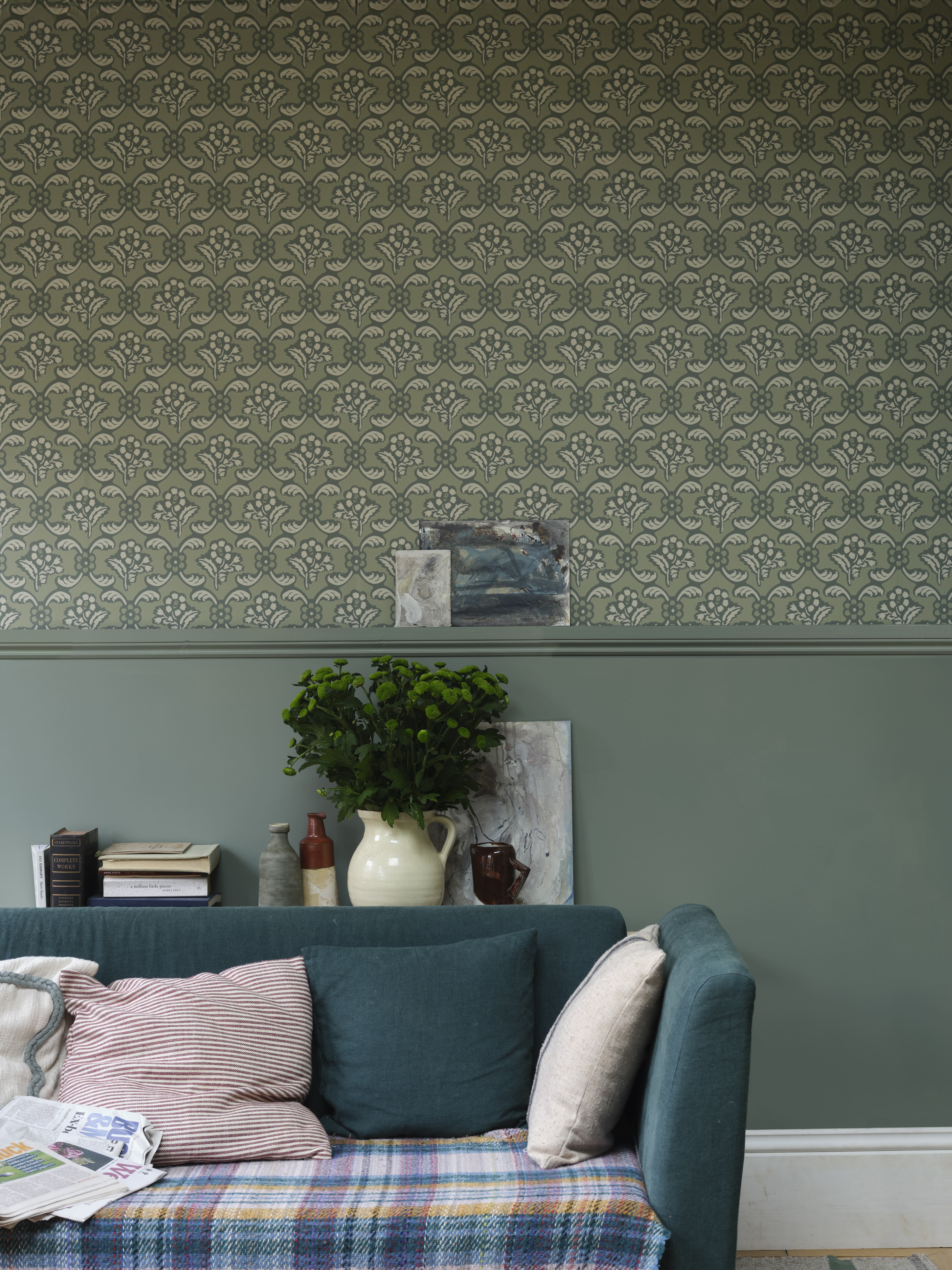 Green living room with patterned green wallpaper and a teal sofa with a plaid blanket. A plant sits prominently in the background.