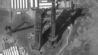 black-and-white satellite photo of a large rocket standing next to a launch tower with dirt and white buildings in the background