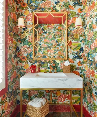 Bold colorful bathroom with bright Chinese inspired wallpaper, rattan mirror, tissue box and basket, wall lights, marble basin vanity unit