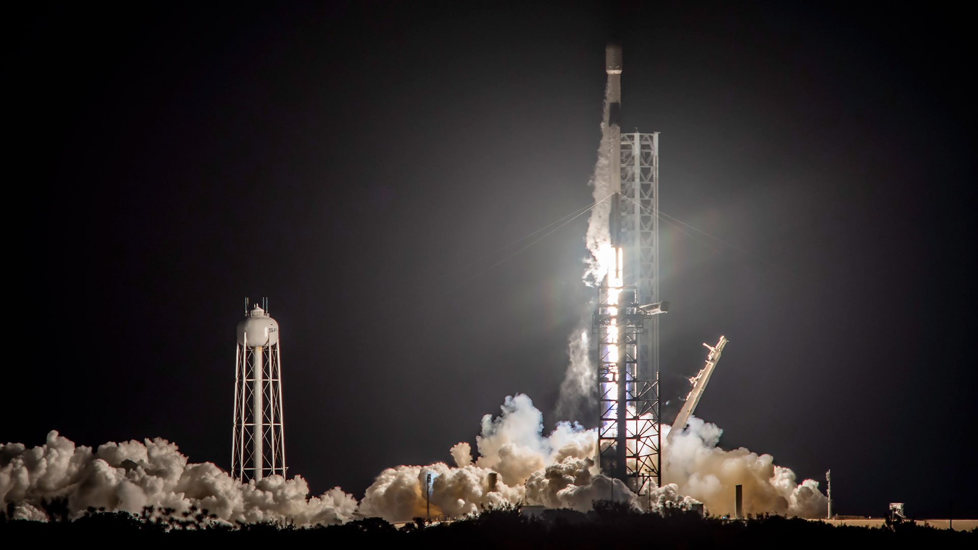 a black and white rocket lifts off from the launch pad into the night sky, lighting the area with its bright white engine plume.