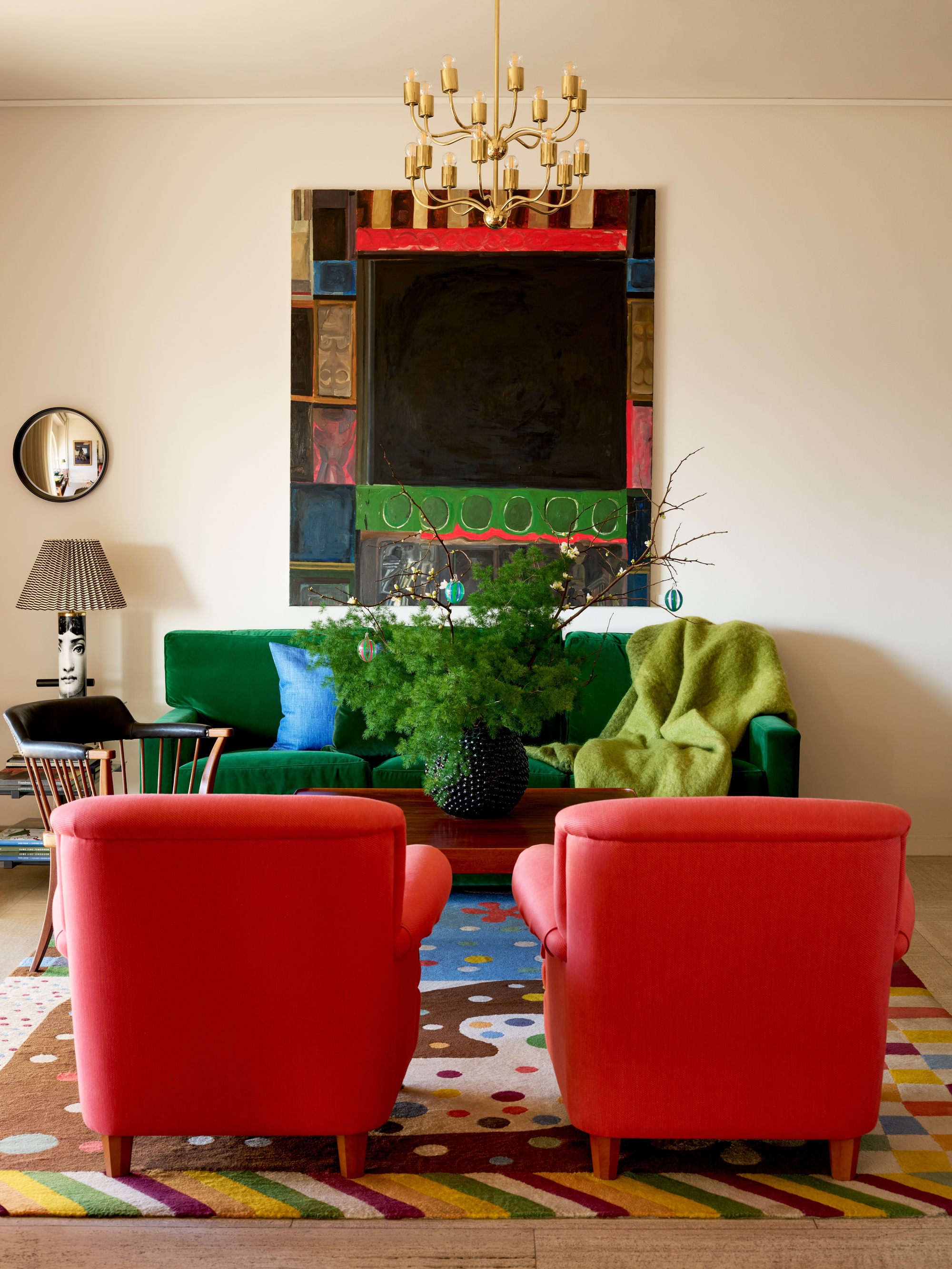 colorful living room with green sofa, two red armchairs, beige walls, bright artwork, ceiling pendant light, side table with lamp and round mirror above it, and colorful patterned rug