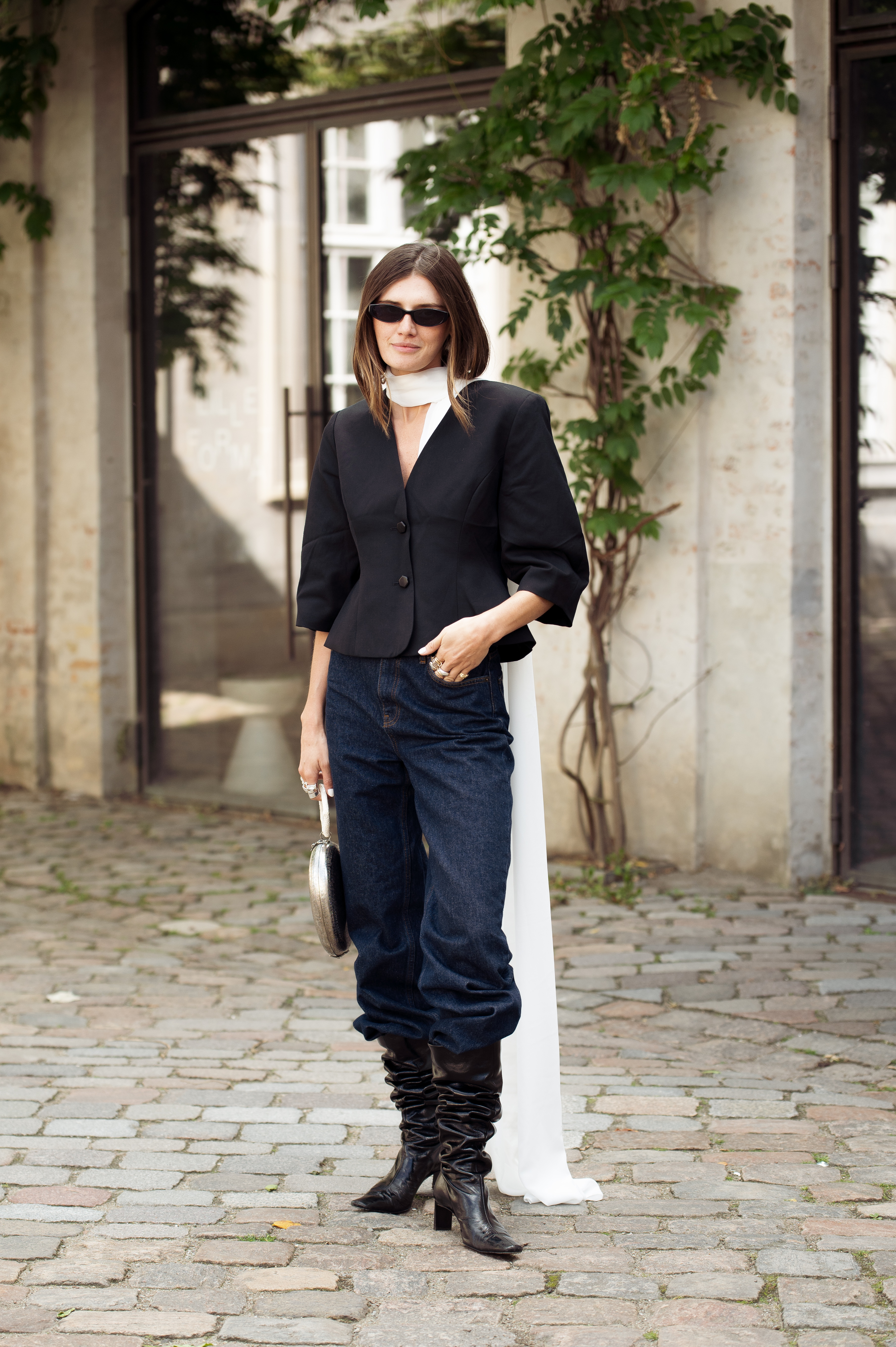 PARIS, FRANCE - JULY 08: A guest wears a short-sleeve white knit top layered with a blue and white boucle tweed vest featuring pearl trim and gold buttons, dark blue straight-leg jeans, black leather ballet flats, oversized black sunglasses, and a long pendant necklace with gold medallions outside the Chanel show during the Haute Couture Fall/Winter 2025/2026 as part of Paris Fashion Week on July 08, 2025 in Paris, France. (Photo by Valentina Frugiuele/Getty Images)