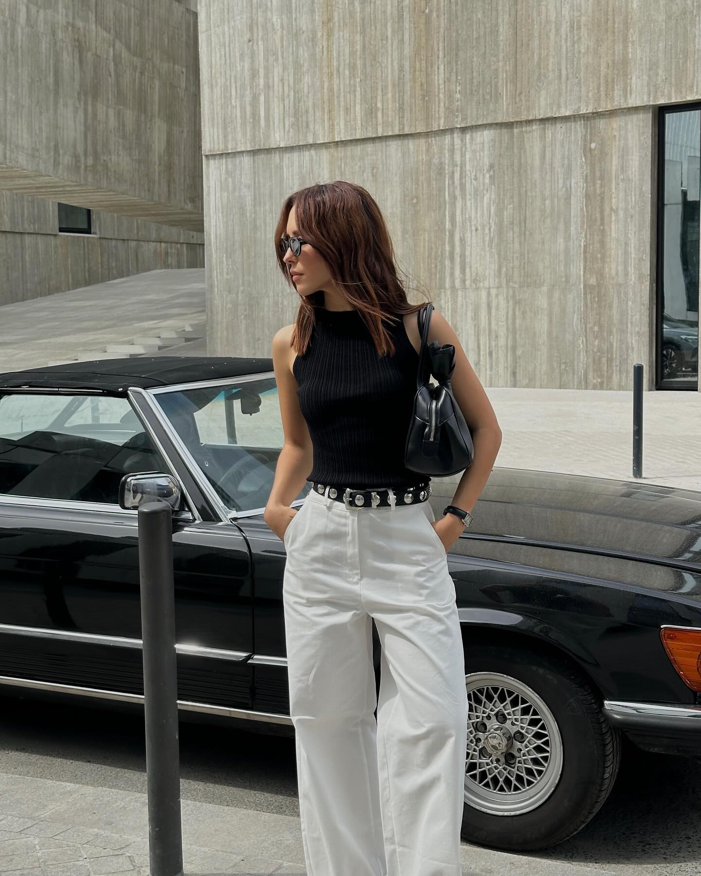fashion influencer Debora Rosa poses in front of a vintage black convertible car wearing black sunglasses, a. black high-neck tank top, a black shoulder bag, a studded Khaite belt, and white casual pants