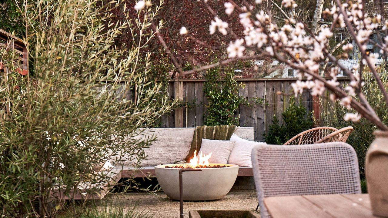 fire pit with garden sofa, cushions and throws, shrubs and tree with blossom, and a table and chair