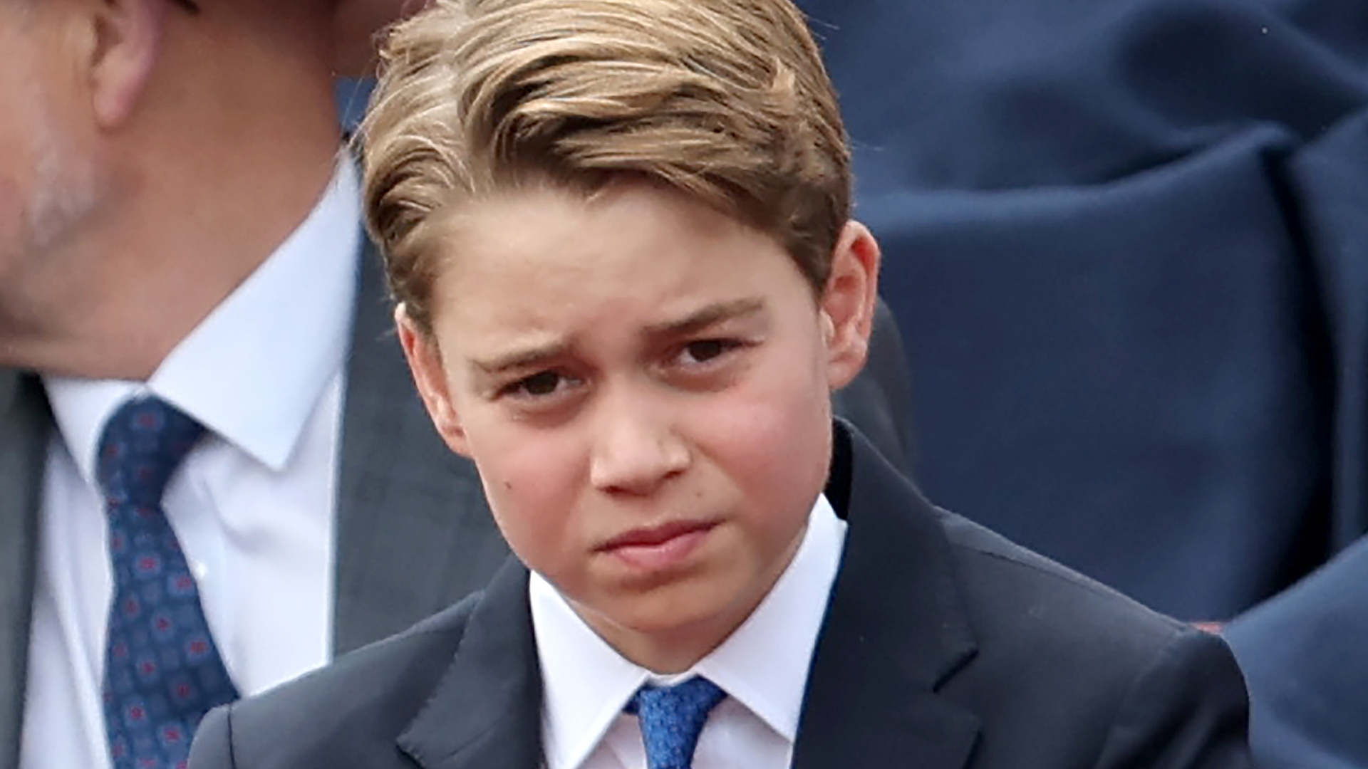 Prince George adjusts his royal blue tie, he is also wearing a navy suit with a white shirt and has blonde brown hair