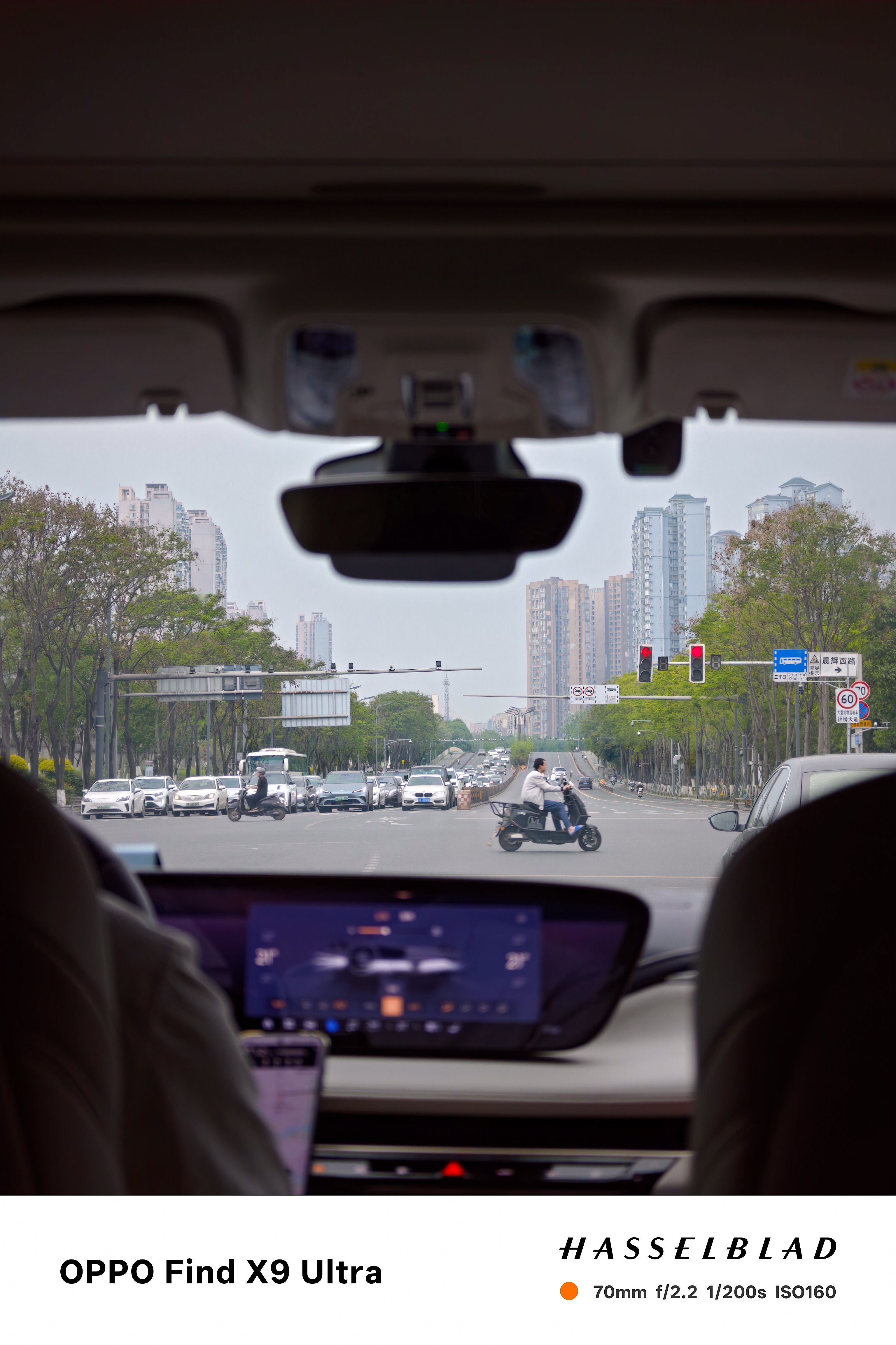 View through a car windshield at a broad city intersection with scooters and traffic