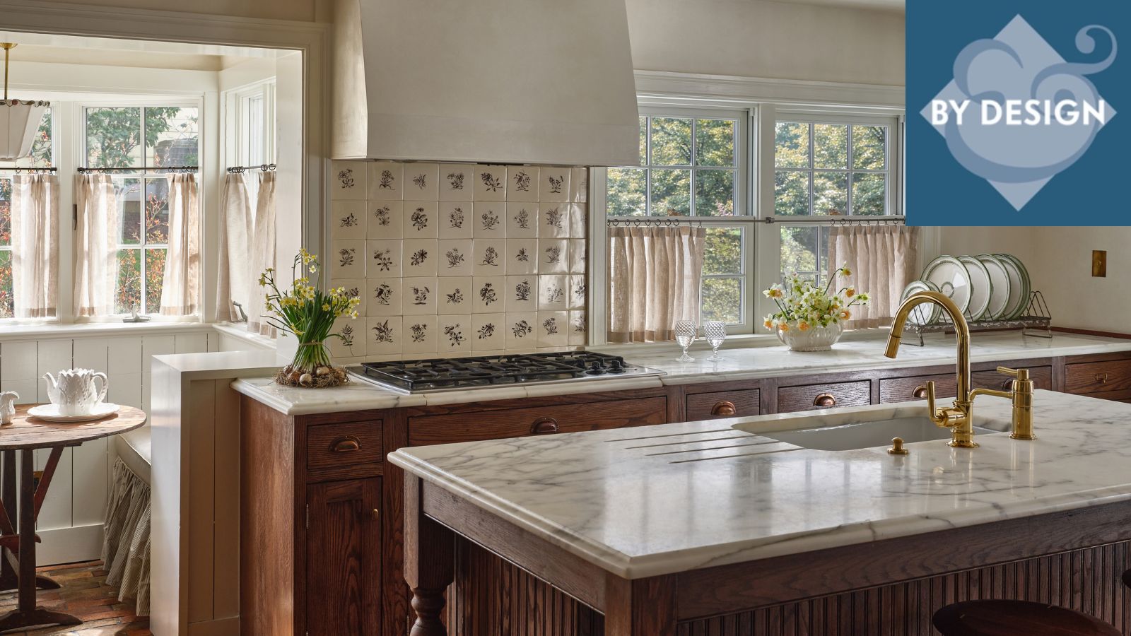 White kitchen with wooden cabinetry and marble countertops. Tiled backsplash with two windows either side with cream cafe curtains. By Design Logo image in top right corner.