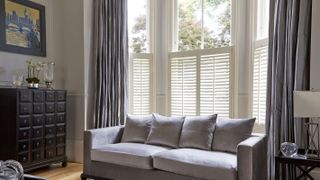 living room with abundant light and high ceilings and large windows, cafe style white sutters and grey curtains and a grey toned sofa