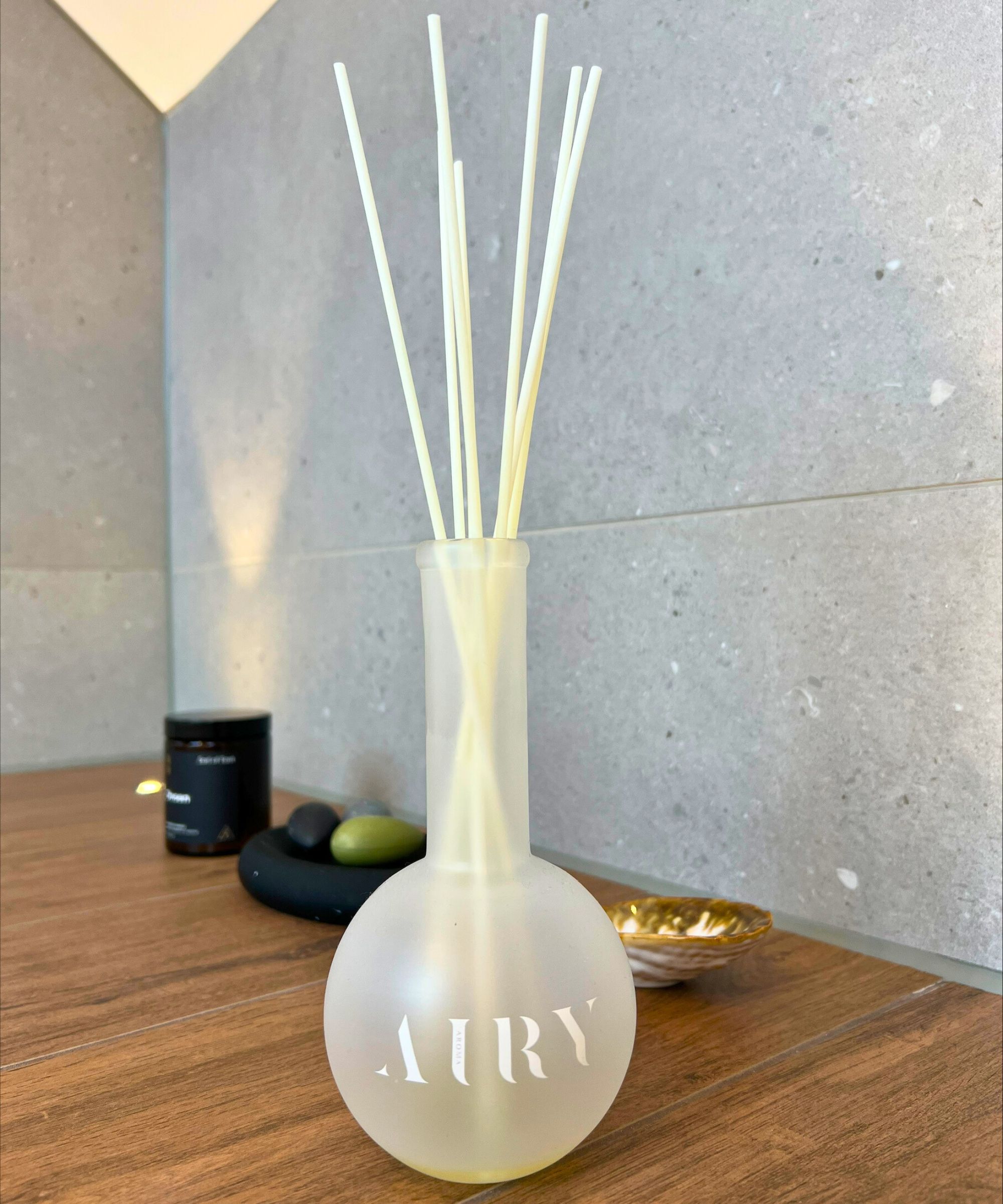 Airy Forest Breath diffuser on a wooden surface with a candle, soaps, and a shell. The walls behind are grey tiled.