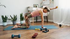 Woman in four-point kneeling position holding dumbbell in front of her, doing Pilates with weights on yoga mat surrounded by small dumbbells and resistance bands