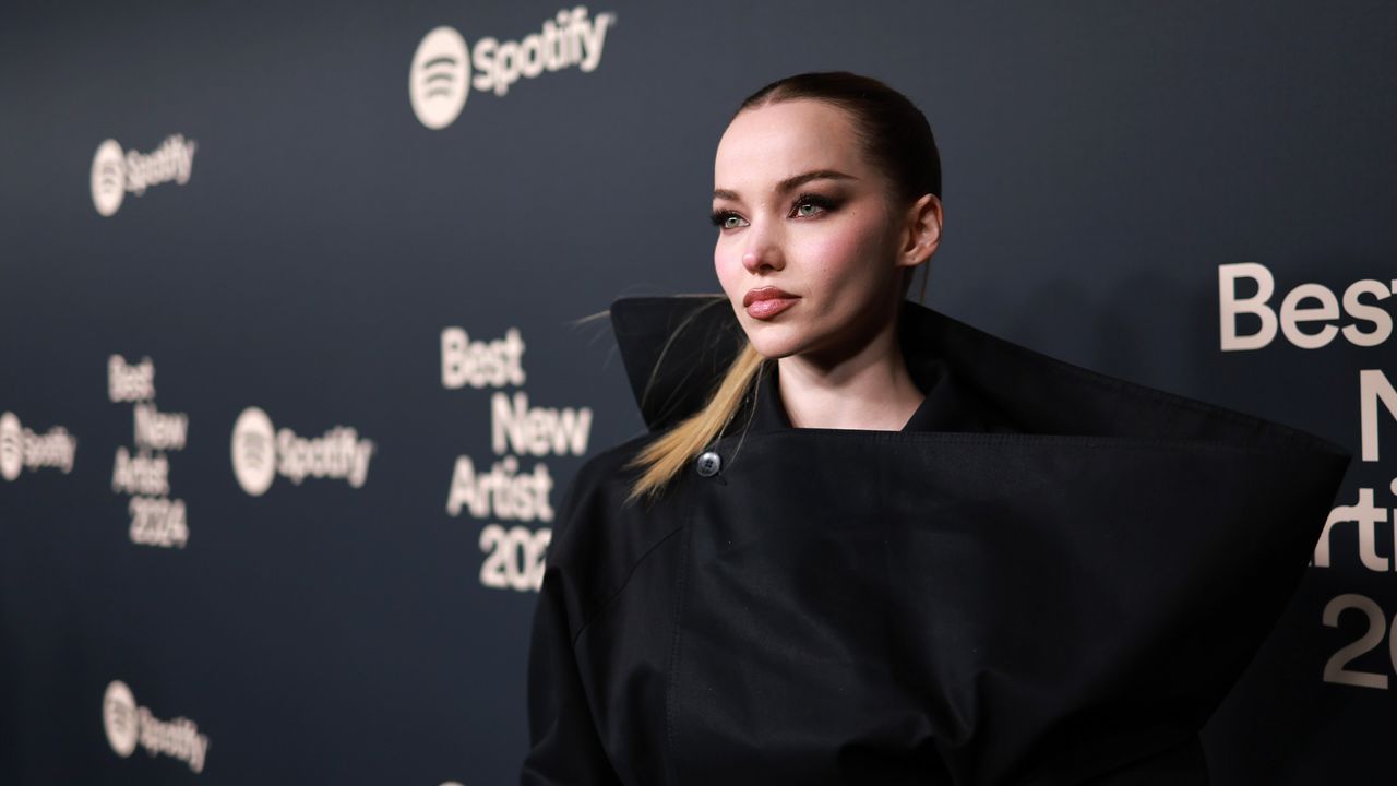 Dove Cameron stands in front of a step and repeat with blonde hair