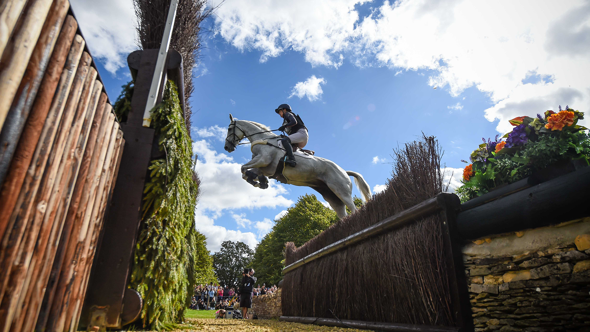 Holly Richardson, a rider in a dark top, rides grey horse Ballyneety Silver Service over a huge ditch and hedge cross-country fence at Burghley