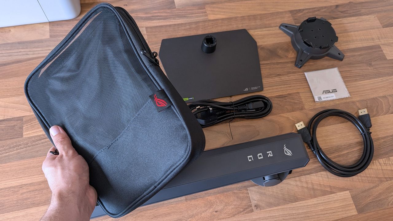 ASUS ROG Strix OLED monitor accessories arranged on a desk, including stand parts, cables, a carrying case, and setup materials.