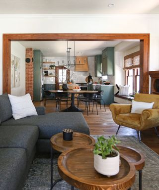 A rustic small living room with a wooden frame leading through to kitchen diner, a gray L-shape couch, warm yellow velvet accent chair, wooden floors and coffee table