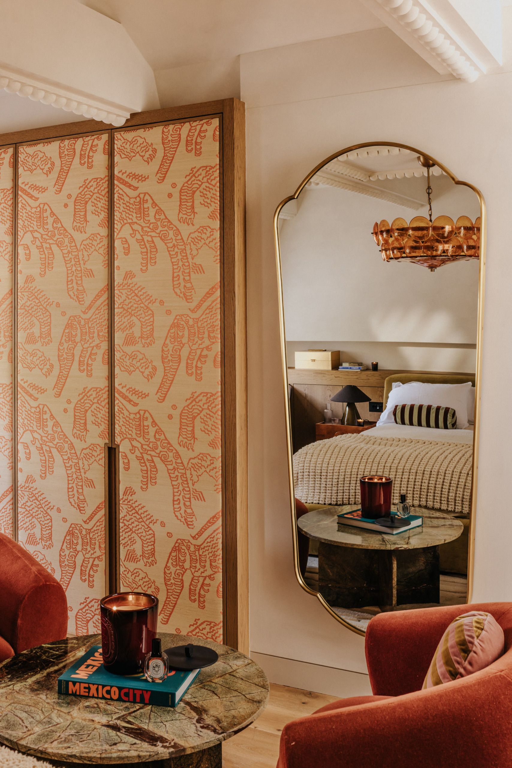 Image of a sitting area in a bedroom with a floor-length mirror, a wardrobe with pink tiger upholstery, two red accent chairs, and a green marble table.