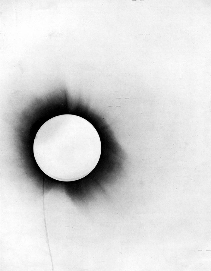 A negative image of the 1919 eclipse plate shows the Sun's disc as a bright white circle surrounded by a dark, radiating corona against a pale background.