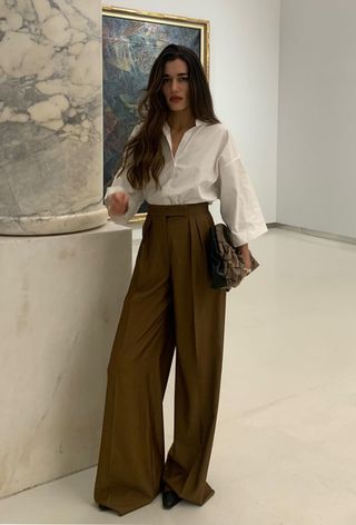 A snakeskin purse is shown in a street-style photo of a woman standing inside an art gallery wearing a white button-down shirt with dark brown wide-leg trousers, brown pointed heels, and a brown snakeskin clutch bag