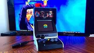 Neo Geo Mini on woodgrain table with gameplay in backdrop on TV.