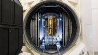a tall cylindrical spacecraft wrapped in gold foil can be seen through a round door in a large metal chamber