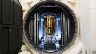 a tall cylindrical spacecraft wrapped in gold foil can be seen through a round door in a large metal chamber