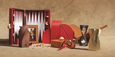 A series of sculpturally designed everyday objects in red, orange, raw wood, gold, and orange, including a Backgammon set, frames, hourglass, and leathery storage units sit on a sand-shaded neutral background.